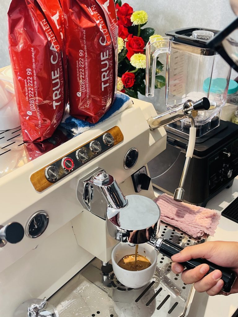 Dòng espresso chảy vào tách từ máy pha, bên cạnh là hai túi cà phê True Cafe