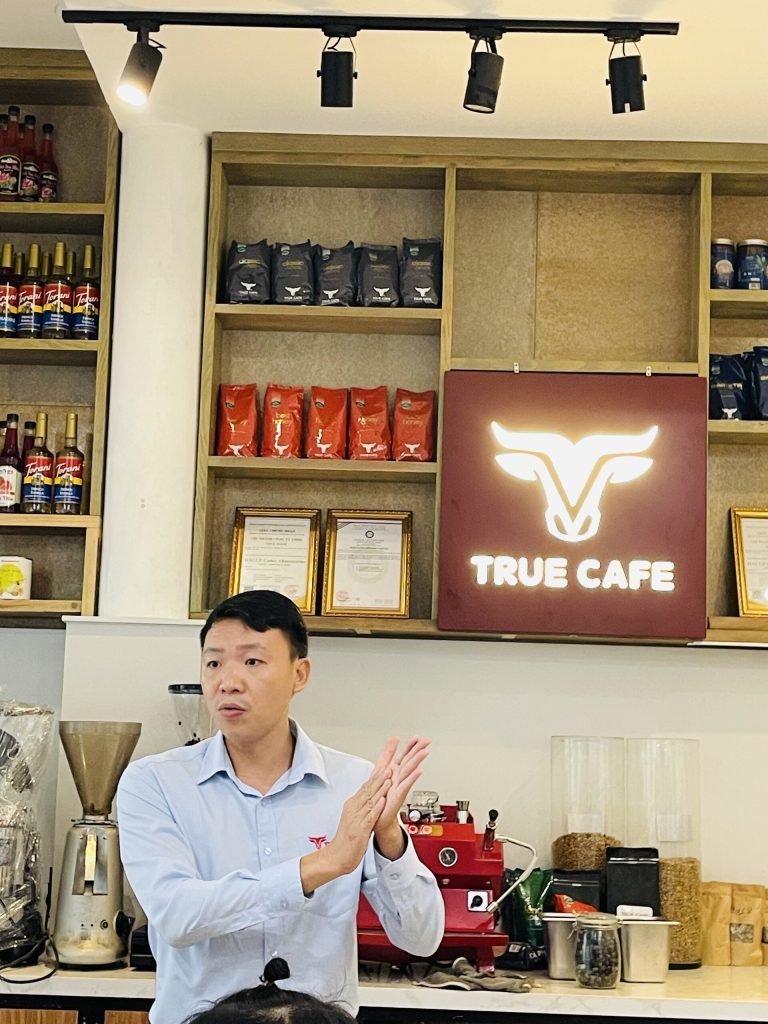 Chuyên viên True Cafe đứng trước quầy bar có logo, hướng dẫn quy trình pha chế