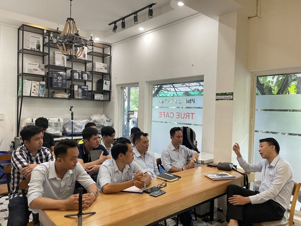 Nhân viên đồng phục True Cafe ngồi bàn dài trong lớp đào tạo, kệ thiết bị phía sau
