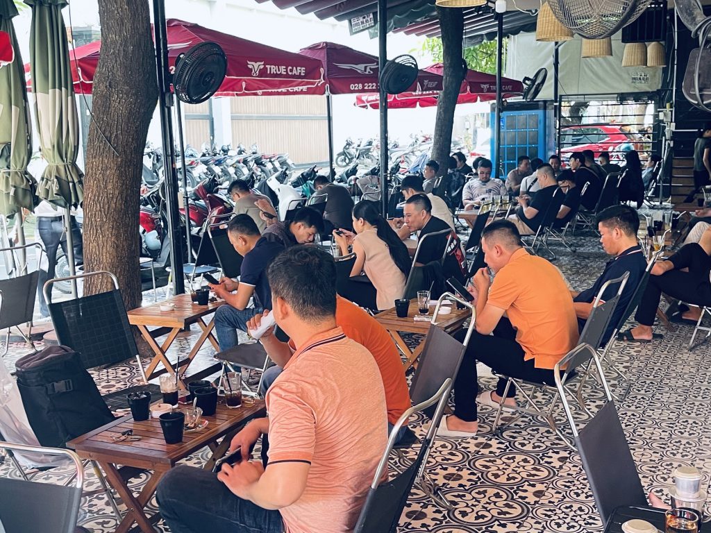 Khách ngồi kín các bàn dưới dàn dù đỏ in logo True Cafe, xe máy đậu dài phía sau