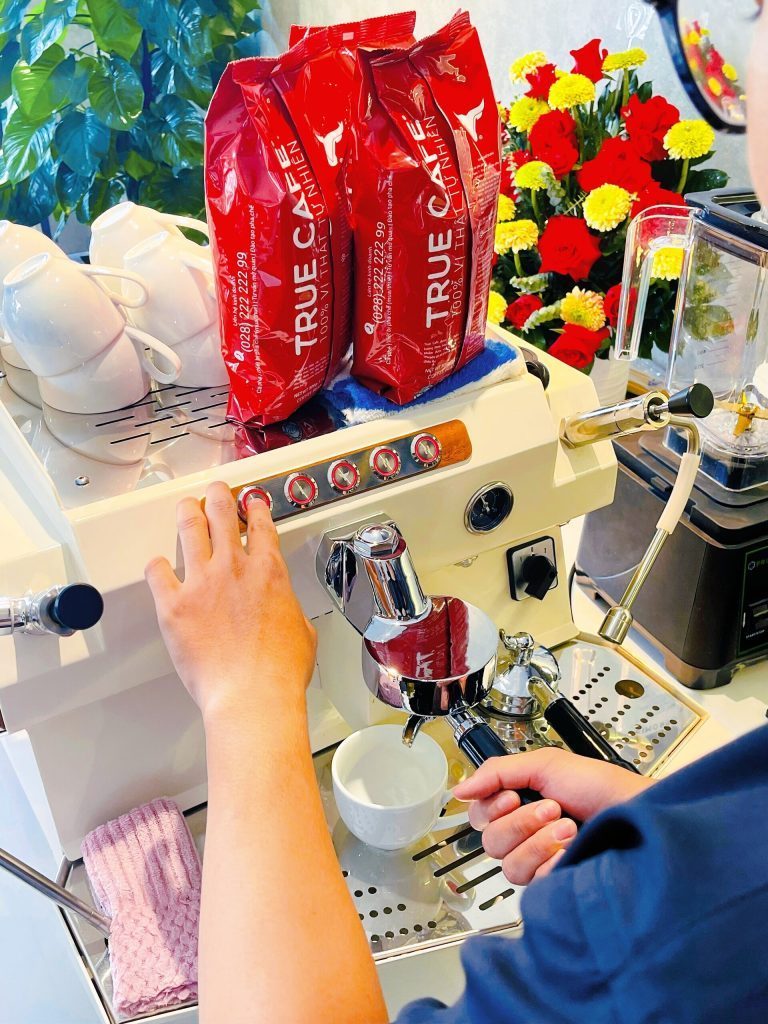 Barista thao tác trên máy pha espresso, bên cạnh là hai túi cà phê True Cafe