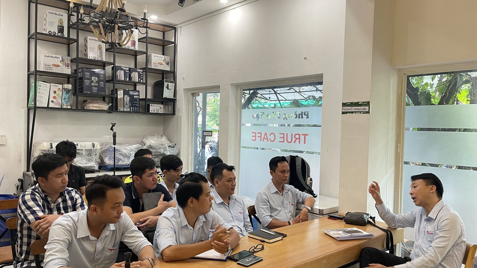 Nhân viên đồng phục True Cafe ngồi bàn dài trong lớp đào tạo, kệ thiết bị pha chế phía sau