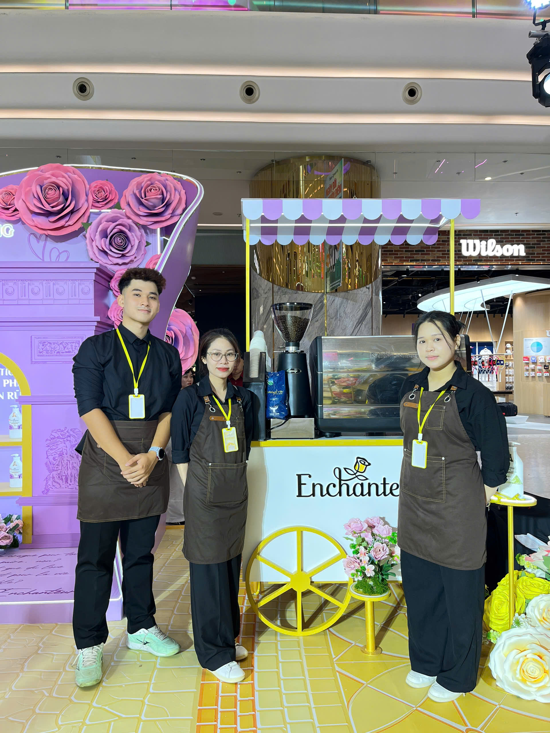 Đội ngũ barista True Cafe đứng cạnh quầy cà phê xe đẩy trang trí theo màu thương hiệu Enchanteur